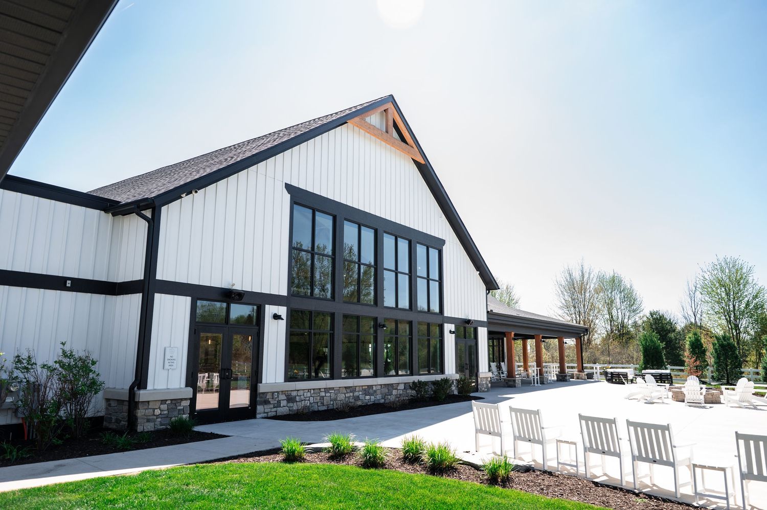 Modern white barn-style event venue with black trim, large windows, stone accents, and outdoor seating on a sunny day.
