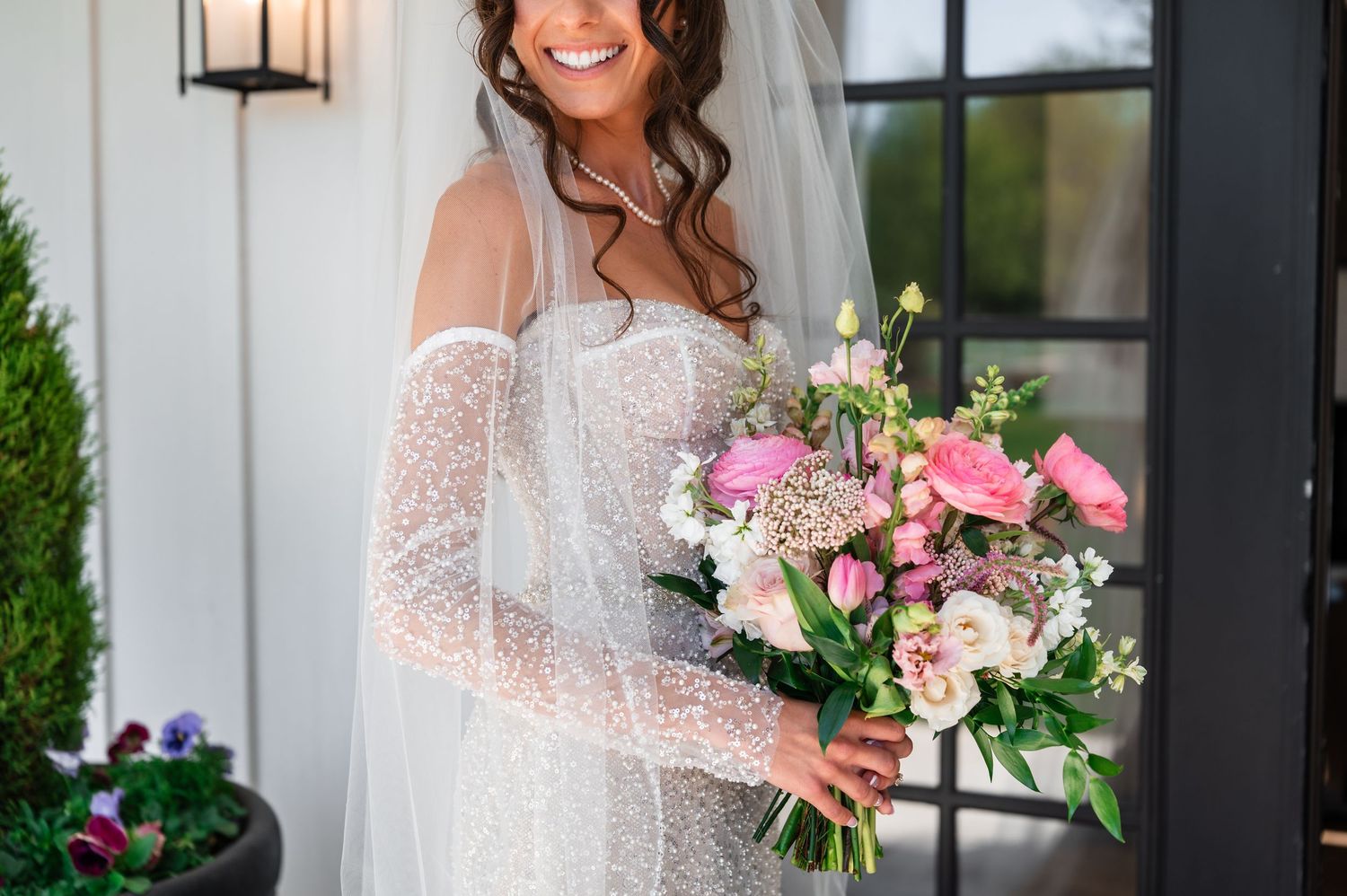 Smiling bride in lace wedding dress and veil holding pink and white floral bouquet outdoors.