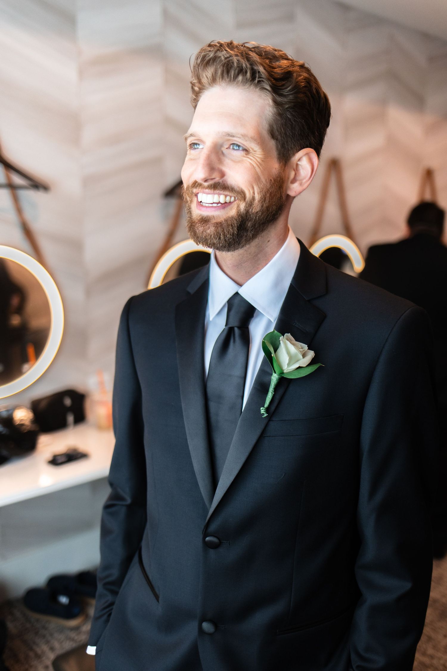 Smiling groom in black tuxedo with green boutonniere, wearing a black tie, standing in a bright modern room.