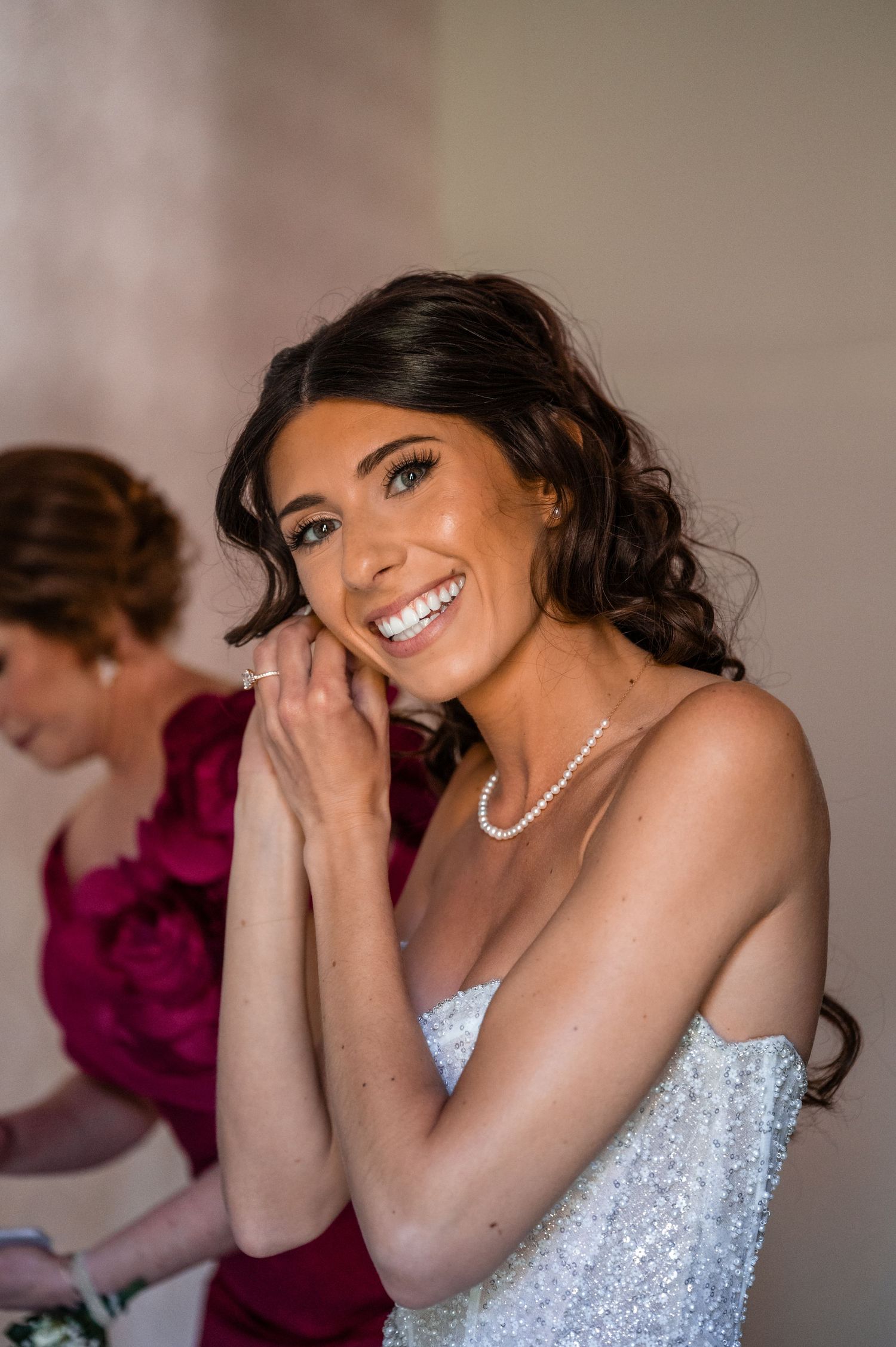 Radiant bride smiling while putting on pearl earrings, wearing a sparkly strapless gown and pearl necklace.