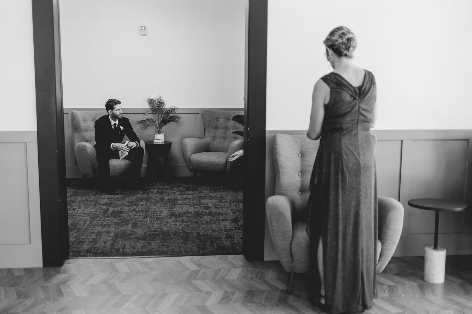 Black and white wedding photo of groom seated on sofa while bridesmaid stands in doorway observing.