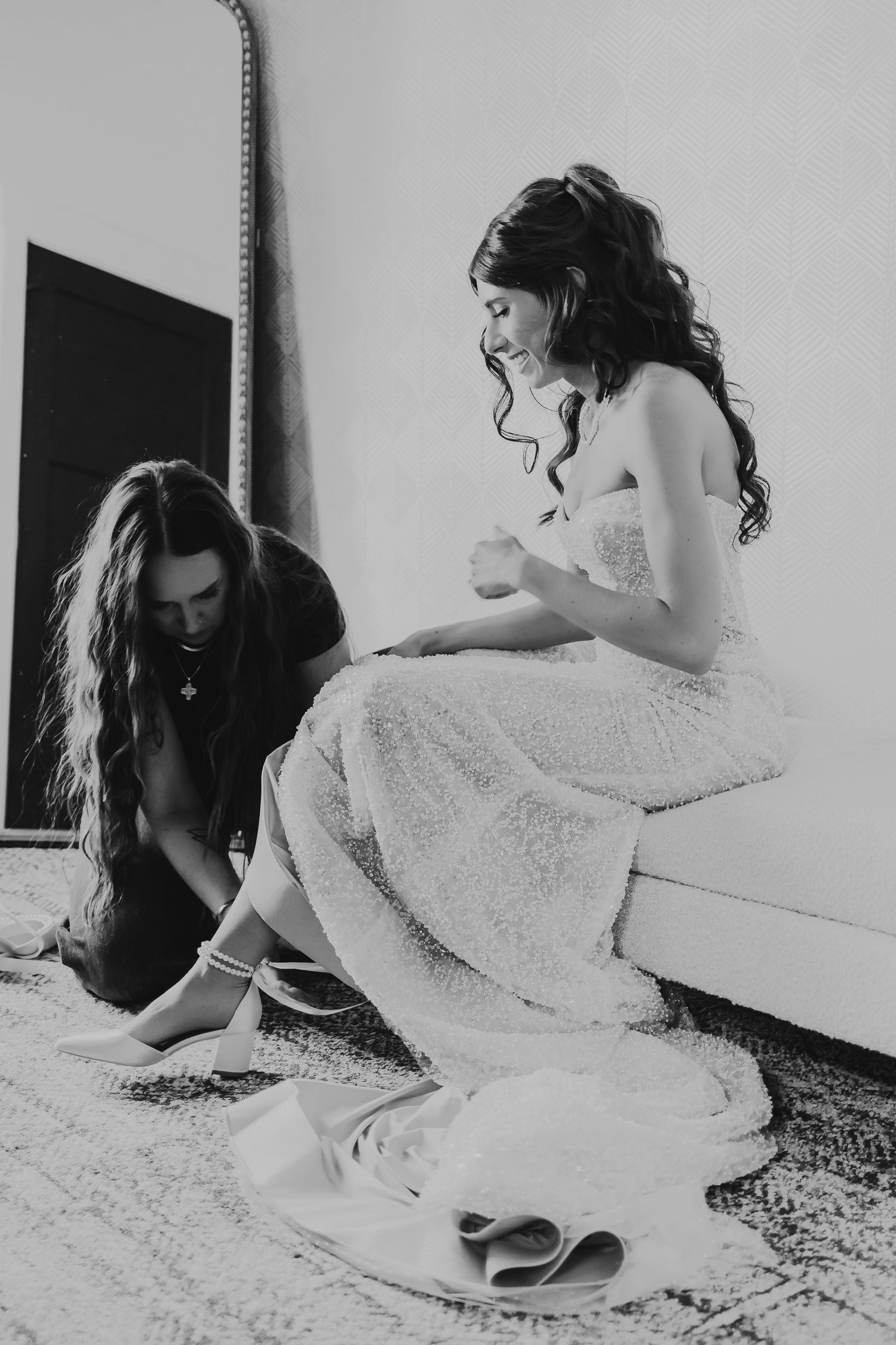 Black and white photo of bridesmaid helping bride with shoes while sitting on bench in wedding dress.