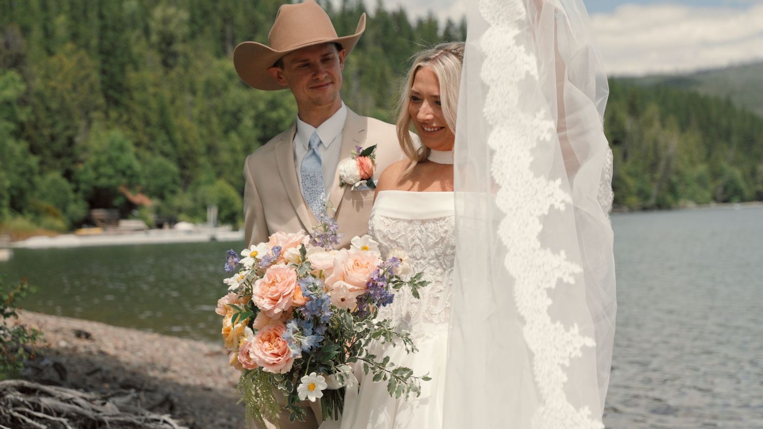 A bride and groom dance joyfully in a sunlit mountain meadow surrounded by evergreen trees on their wedding day.