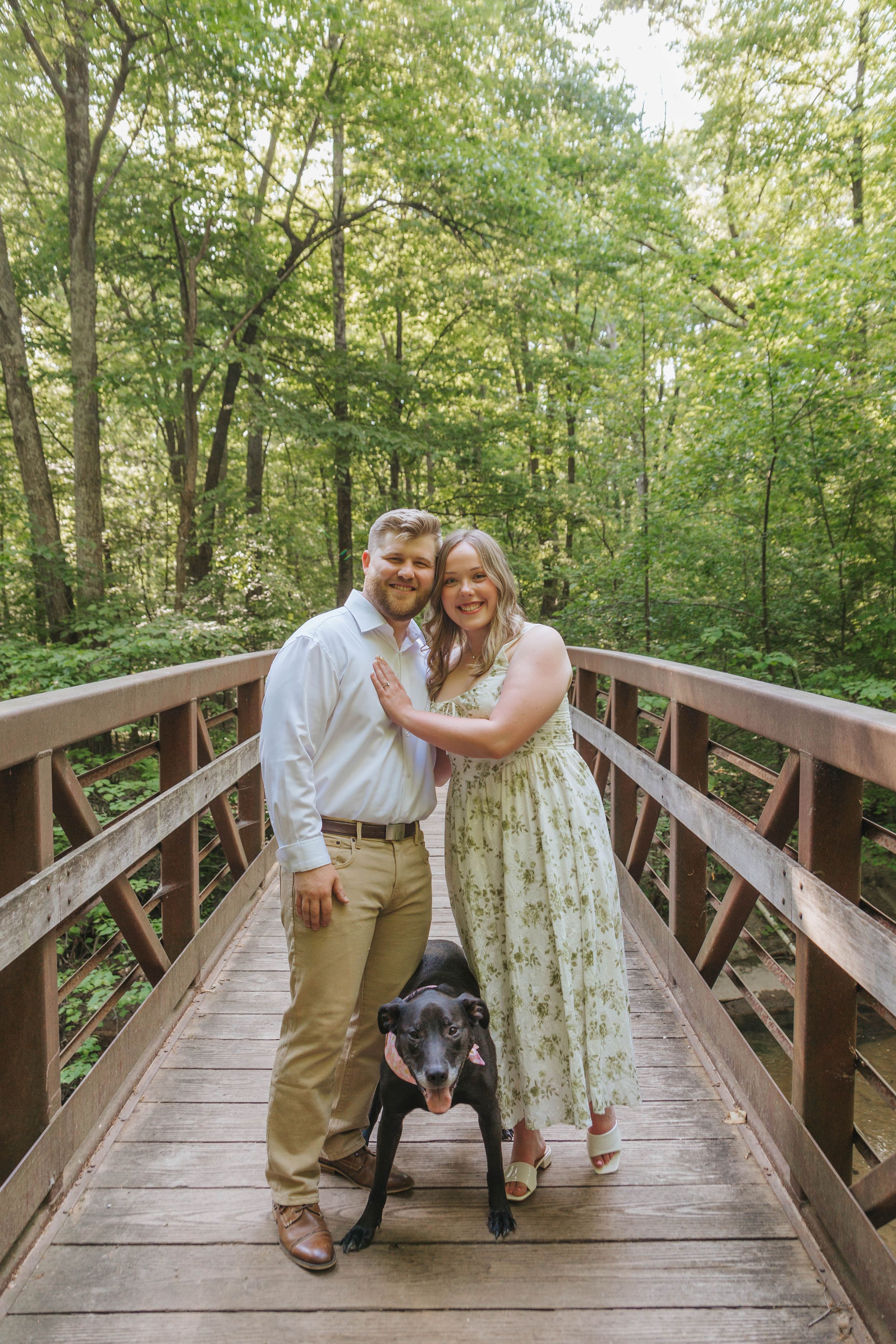 Couple smiling on a wooden bridge in a lush green forest, with a black Labrador dog standing in front of them.