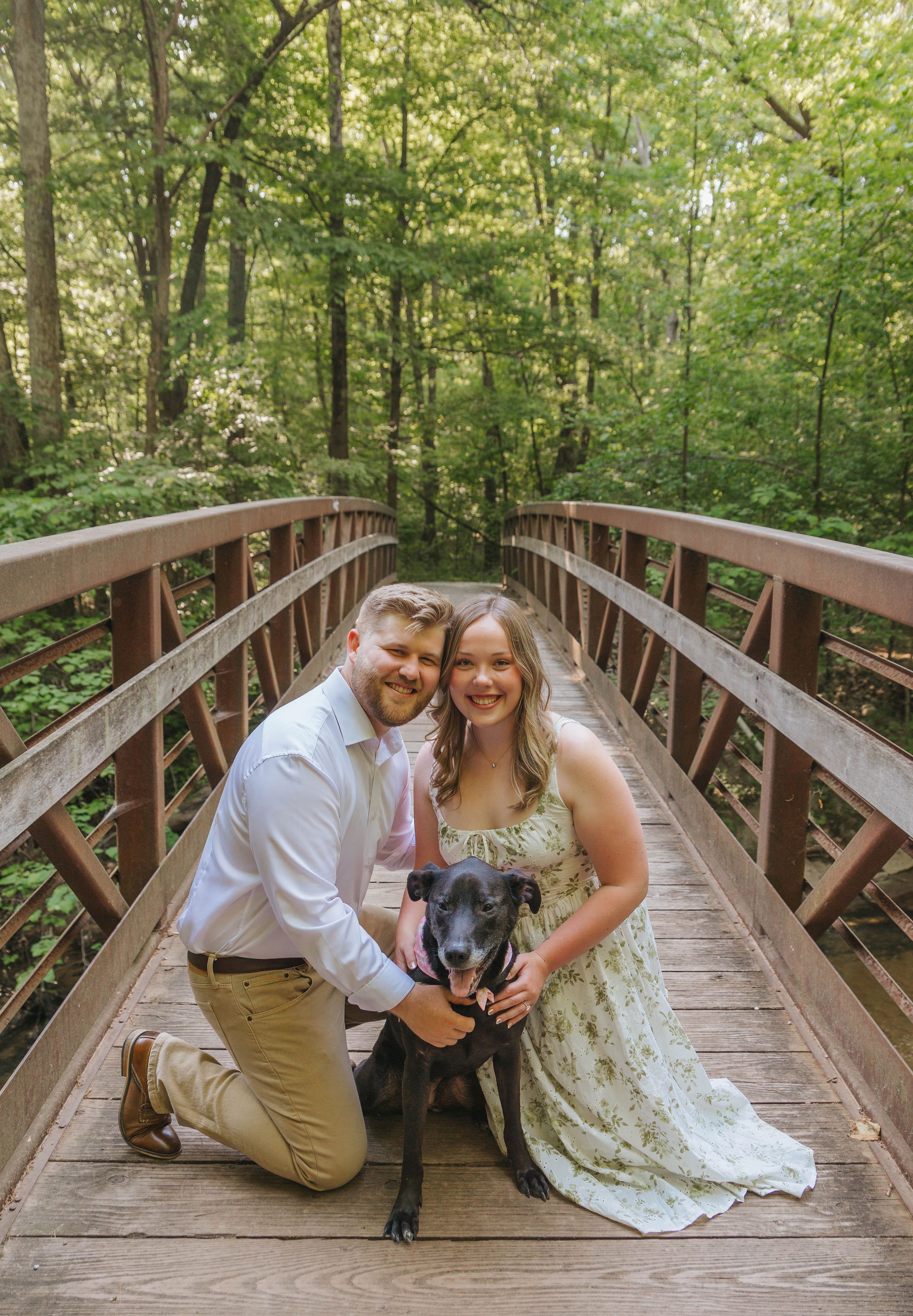 Couple kneeling on a wooden bridge with their black dog, surrounded by lush green forest trees.