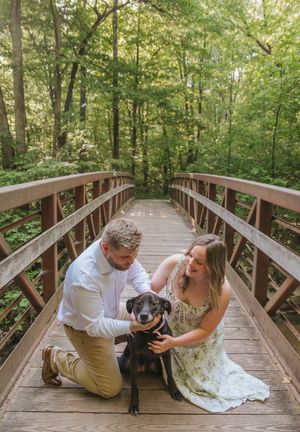 Couple kneeling on a wooden bridge in a lush forest, petting their black dog during an engagement photo session.