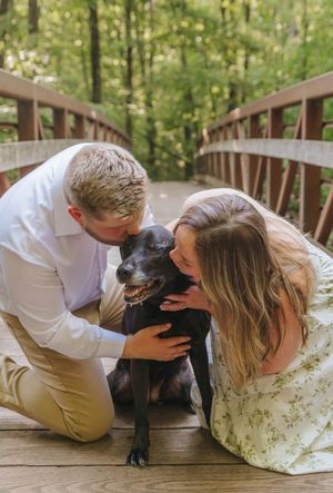 A couple kneels on a wooden bridge, lovingly nuzzling their black dog surrounded by lush green trees.