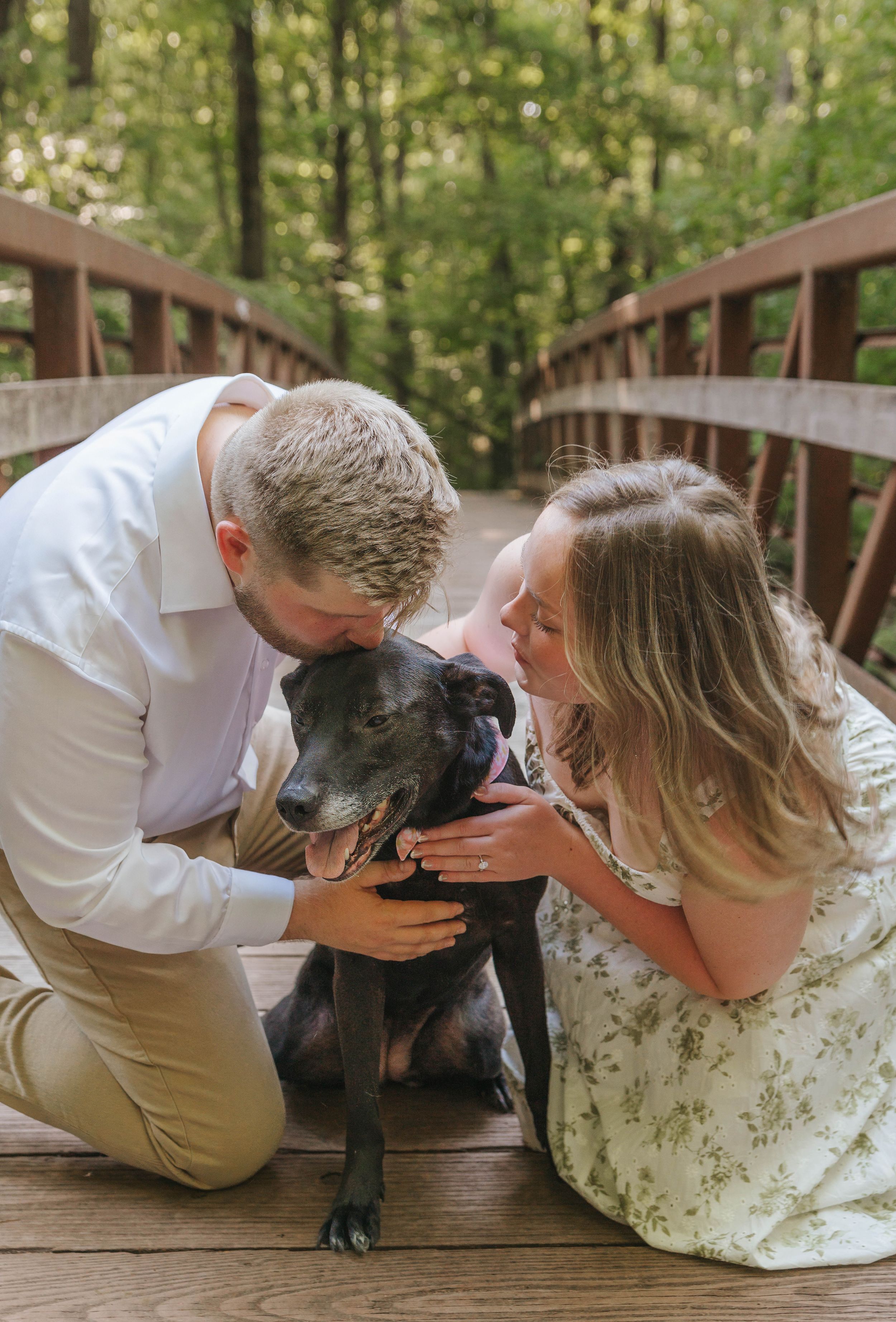 Couple kneeling on a wooden bridge, kissing their black dog, surrounded by lush green trees.