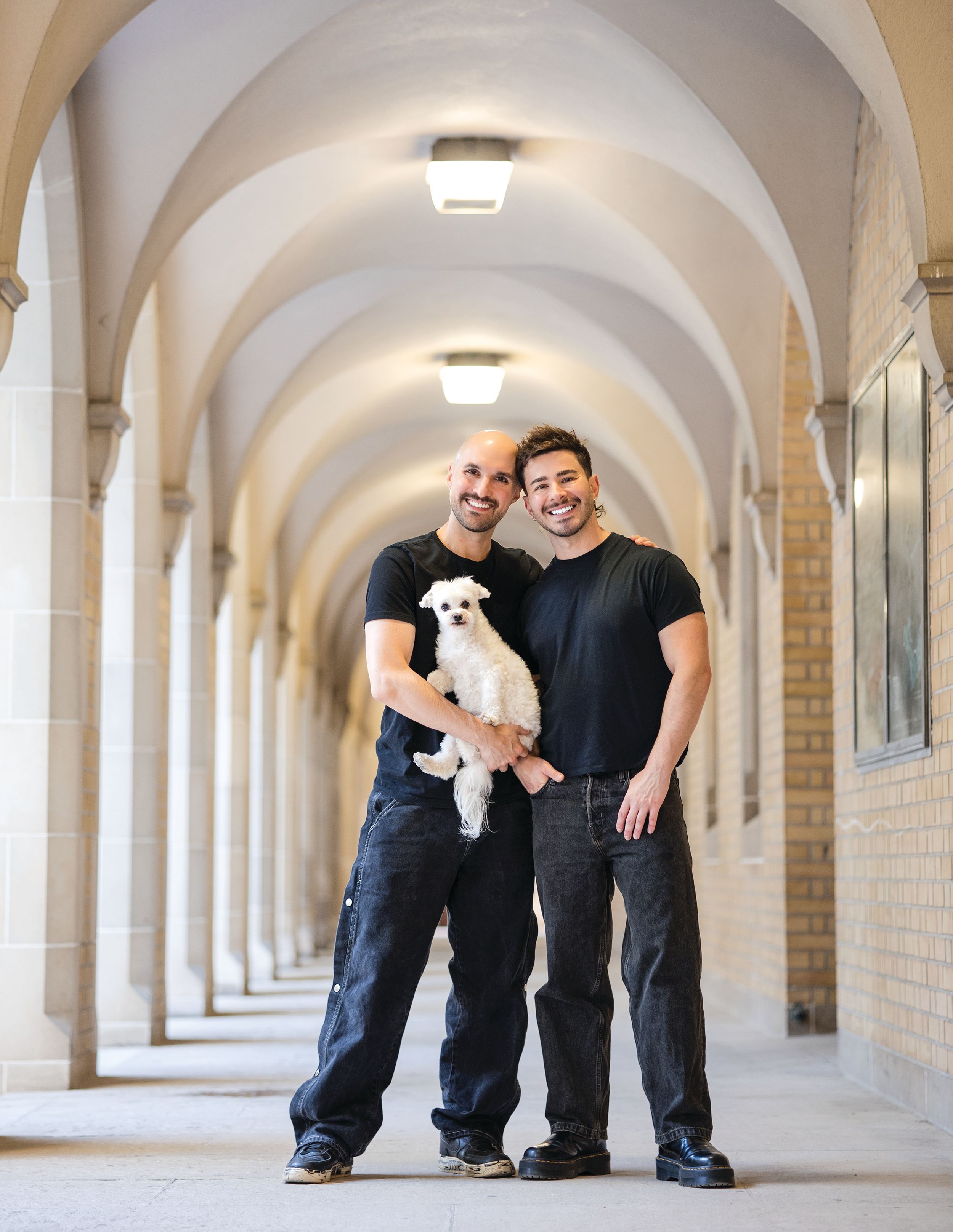 Two men in black outfits hold a small white dog in an arched hallway.