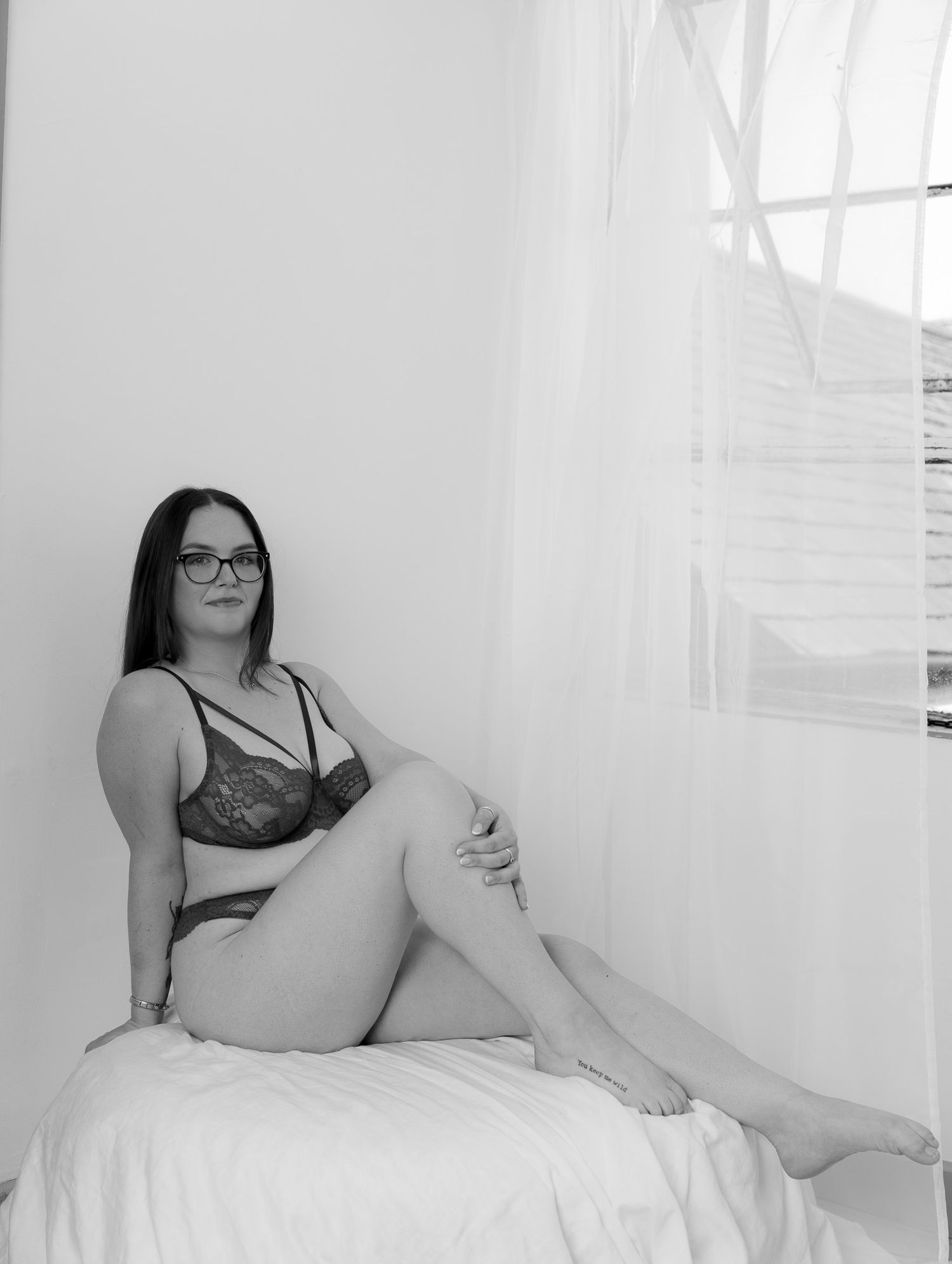 Black and white boudoir photo of a woman in lace lingerie sitting on a bed near a window with sheer curtains.