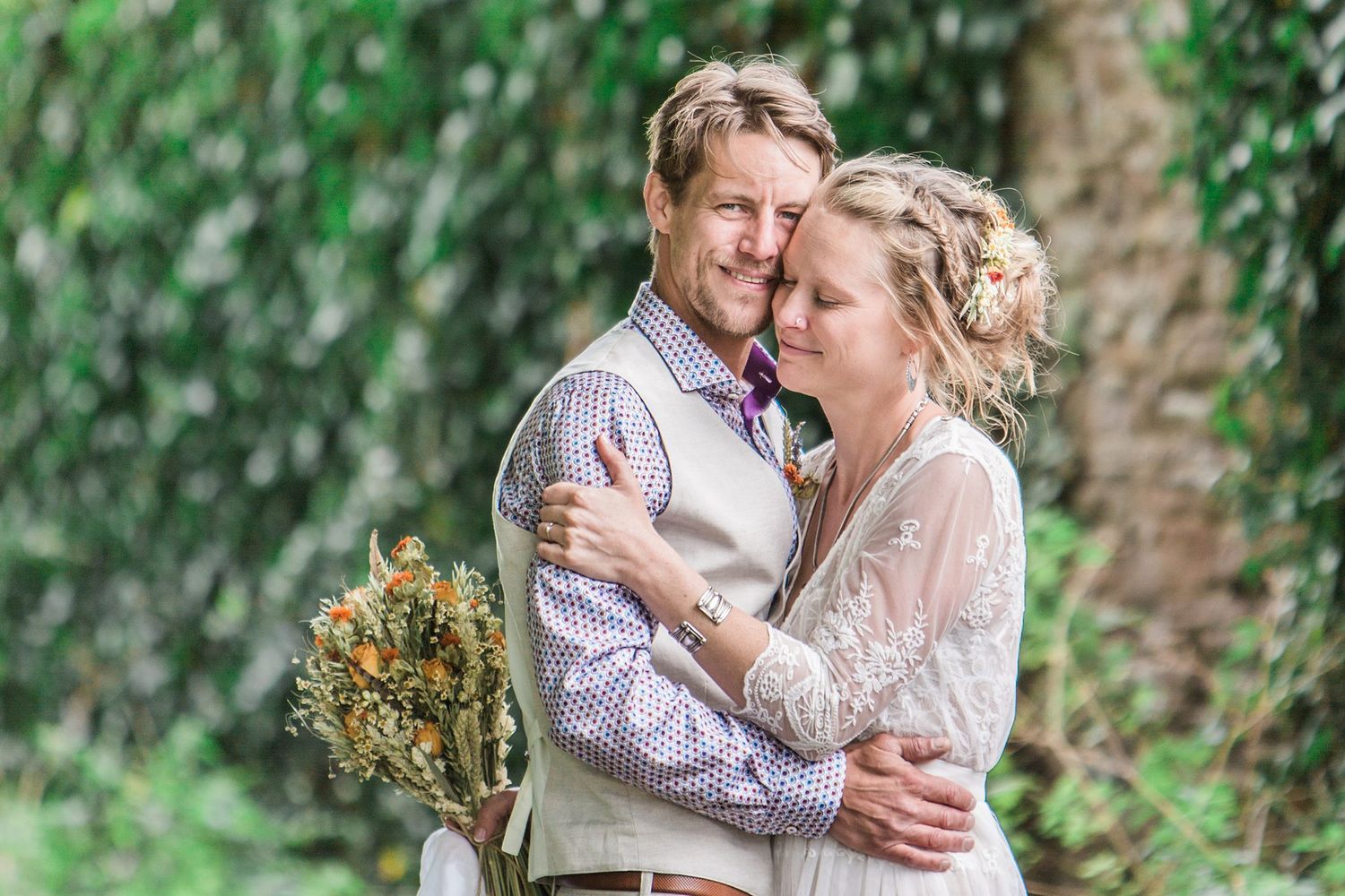 A bride and groom embrace outdoors, surrounded by lush green ivy, as she holds a wildflower bouquet in a romantic wedding portrait.