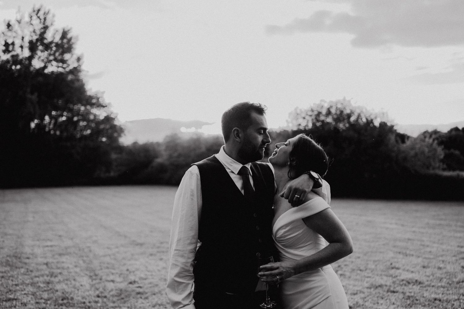 Black and white photo of a bride and groom sharing a romantic moment in an open field at sunset.