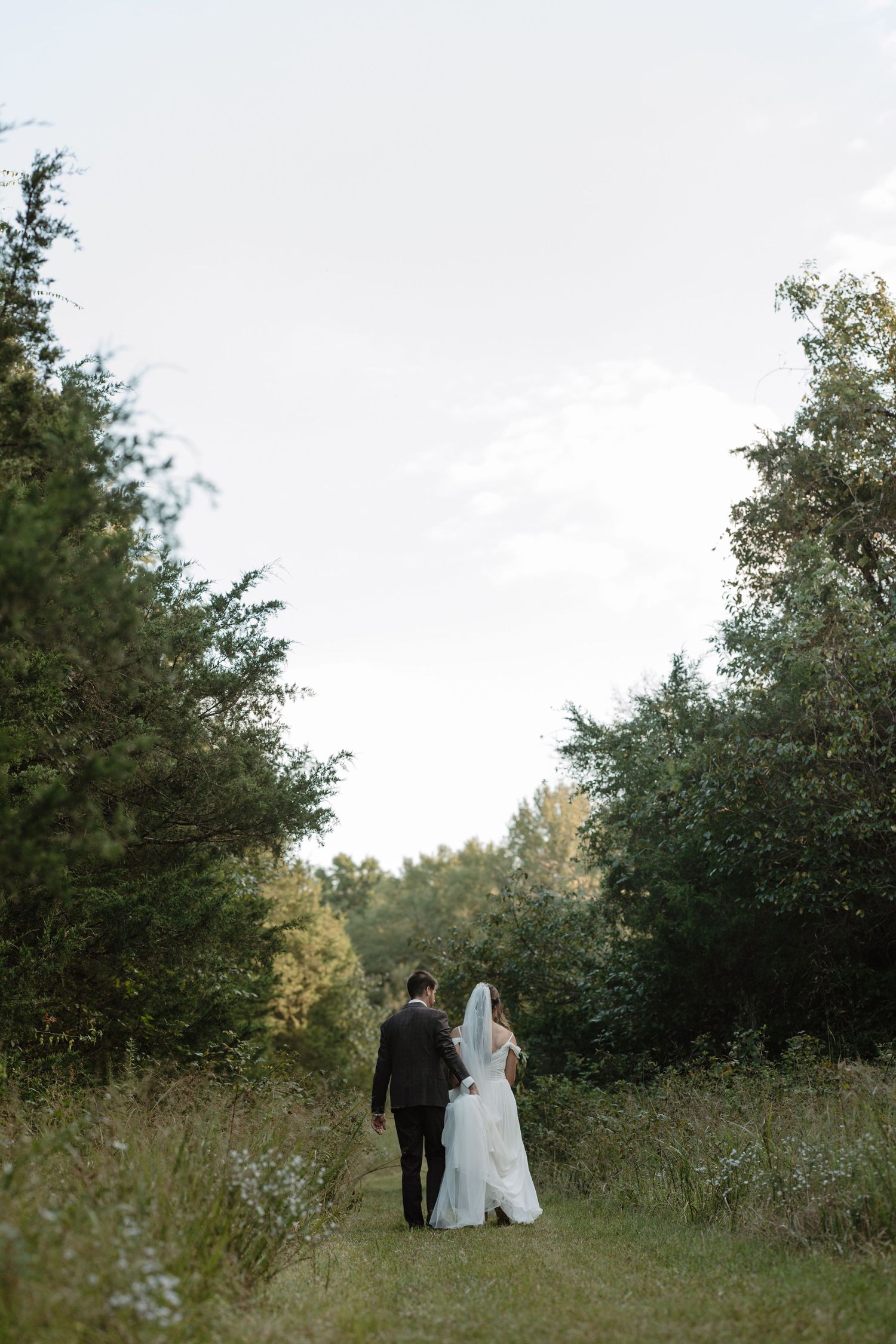 Bride and groom walking hand in hand through lush green tree-lined pathway.