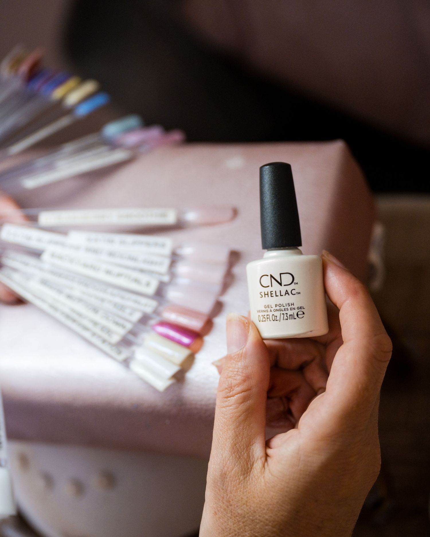 Hand holding CND Shellac gel polish bottle in white, with nail color swatches displayed on pink box in background.