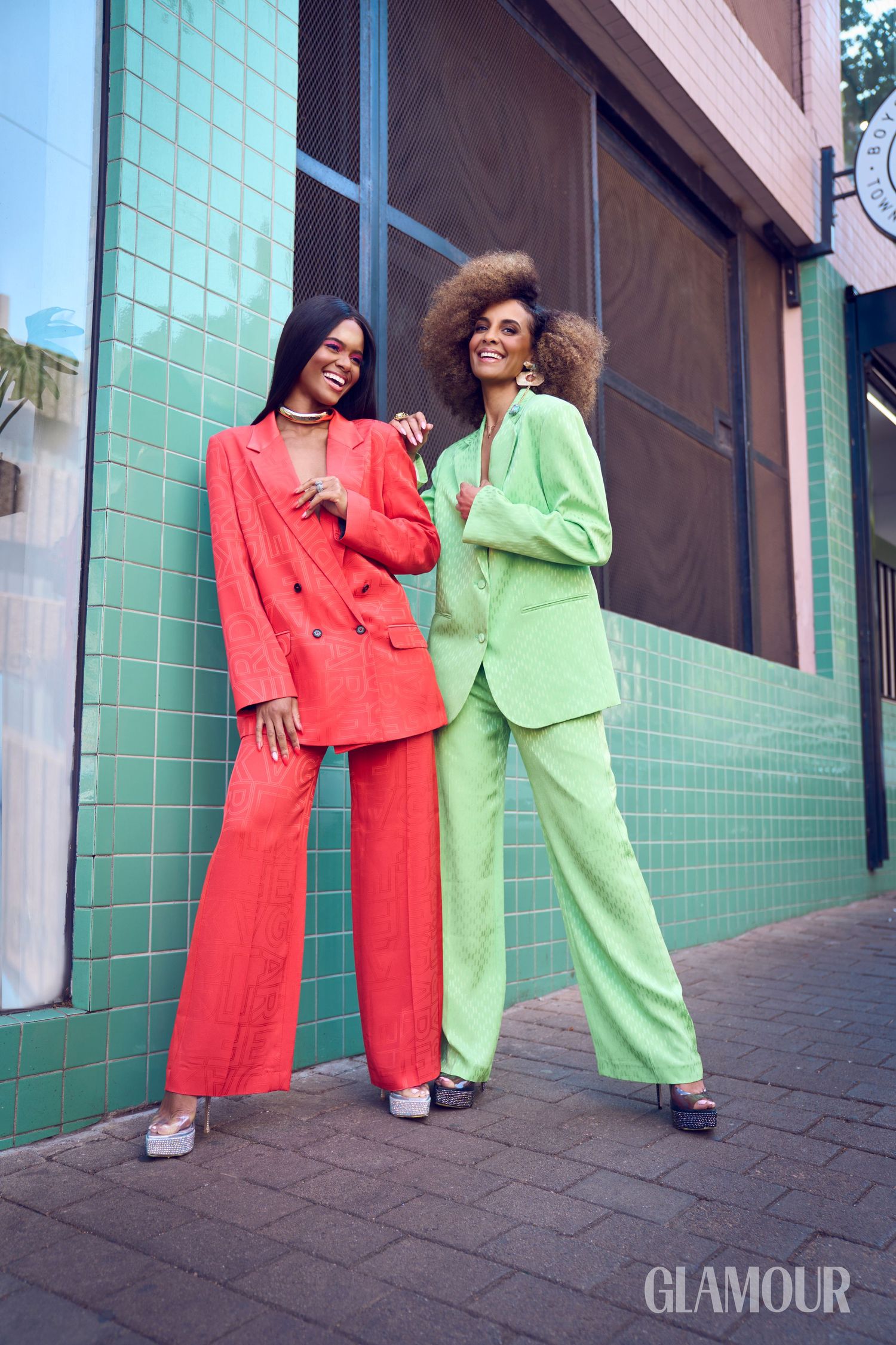 Two women in bold red and green oversized suits pose against a teal tiled wall outdoors.