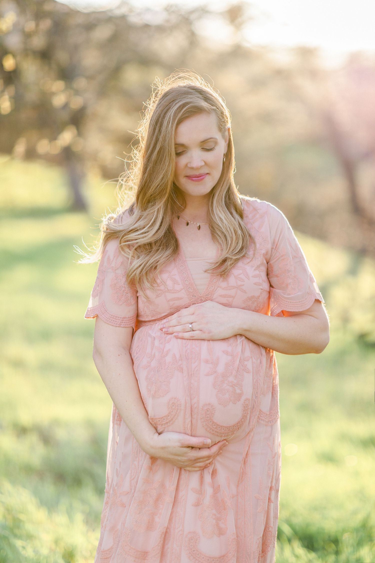 Pregnant woman in pink lace dress cradling baby bump outdoors in sunlit garden during golden hour.