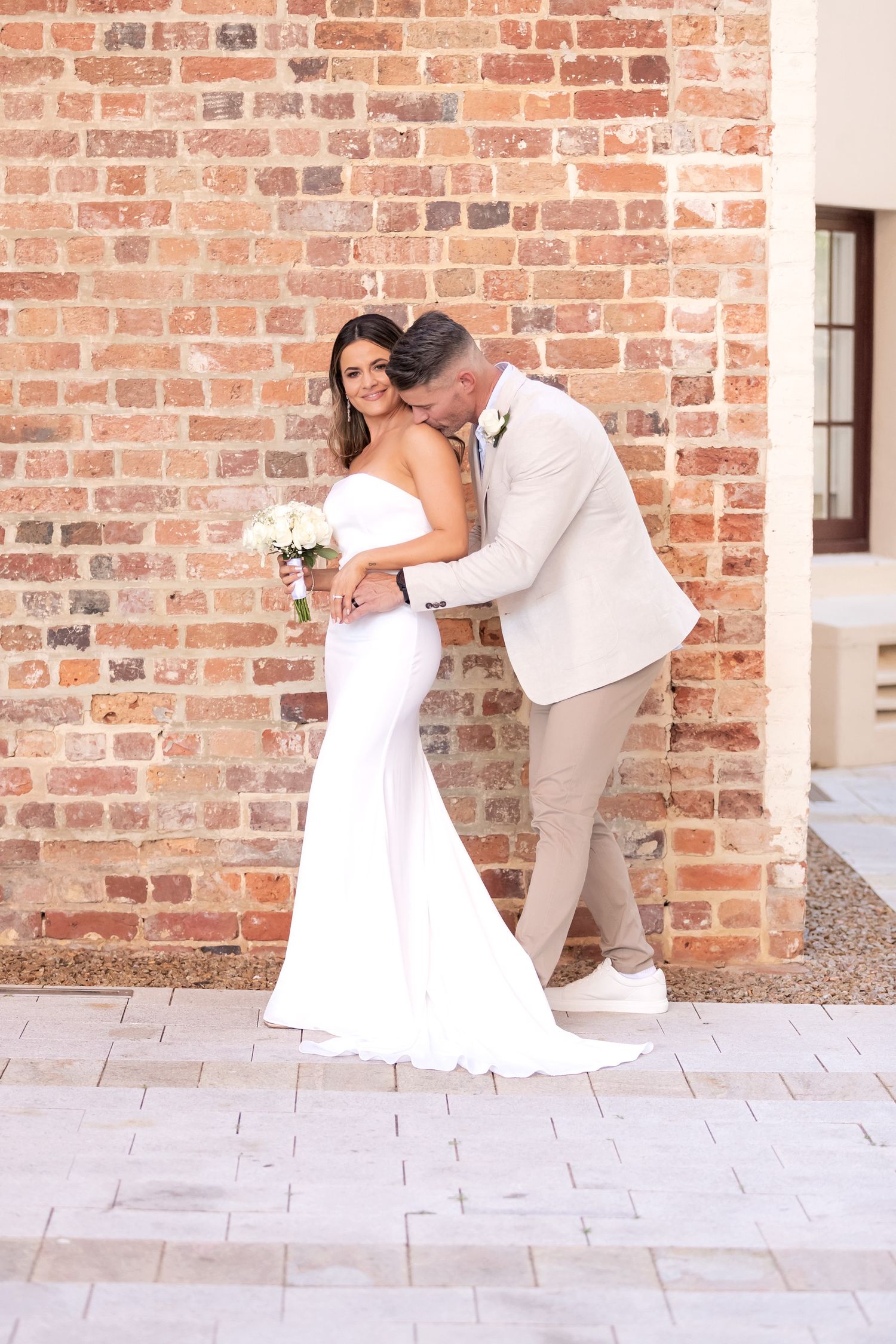 Bride in white gown holds bouquet as groom in tan suit kisses her cheek against a rustic brick wall.