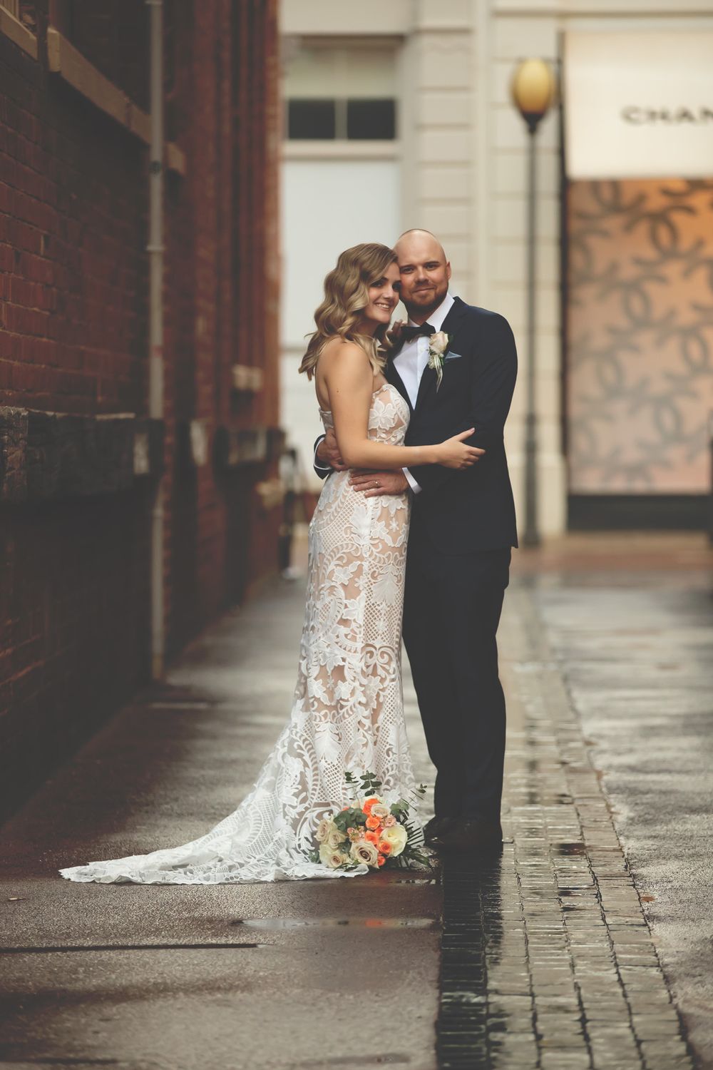 Bride in lace gown and groom in tuxedo embrace on a rain-slicked urban alley on their wedding day.