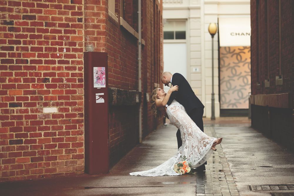 A groom dips his bride in a romantic kiss in a charming brick alleyway, her lace wedding dress flowing gracefully.