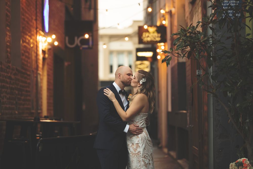 Bride and groom share a romantic kiss in a warmly lit brick alleyway adorned with string lights.
