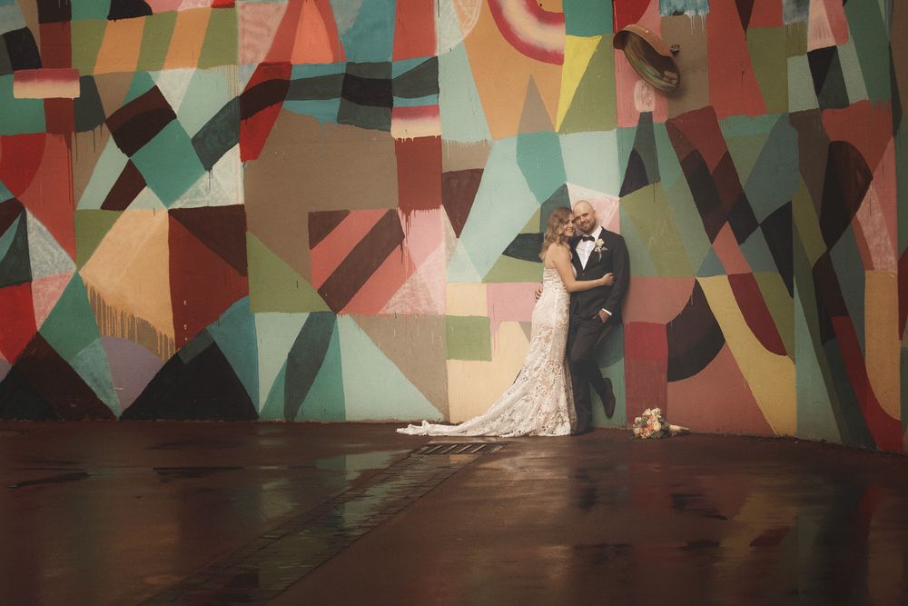 Bride and groom share a romantic kiss in front of a vibrant colorful geometric mural at their wedding venue.