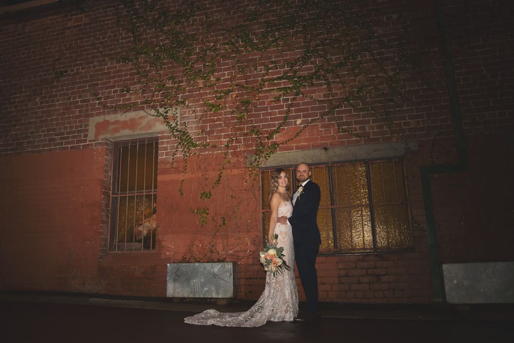 Bride and groom pose romantically against a rustic red brick wall at night, surrounded by ivy and warm lighting.