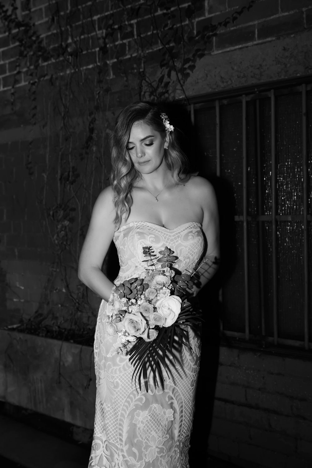 Black and white photo of a bride in a strapless lace gown holding a floral bouquet, standing against a brick wall.