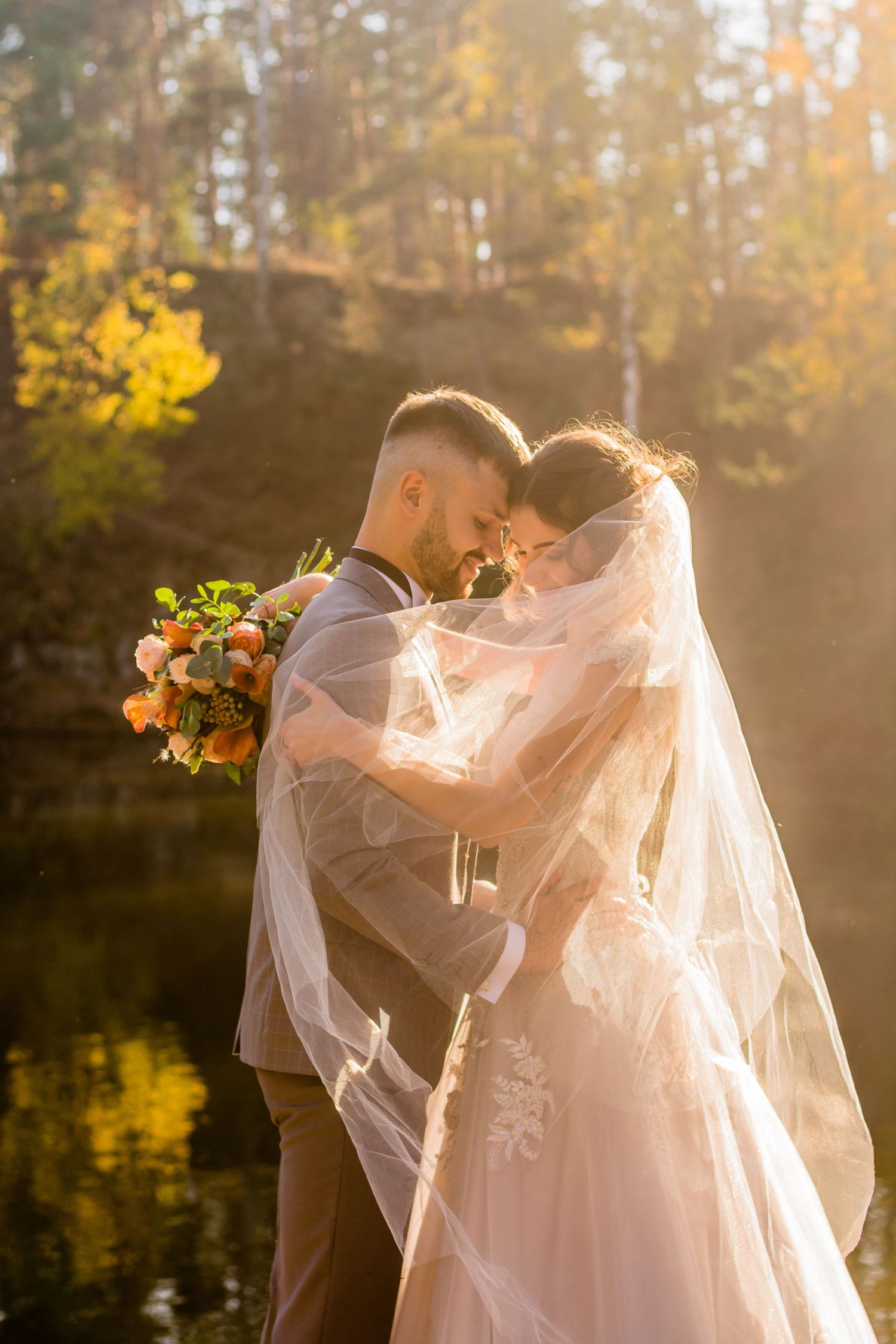 Bride and groom sharing a romantic moment under a veil in a sunlit autumn forest by a reflective pond.