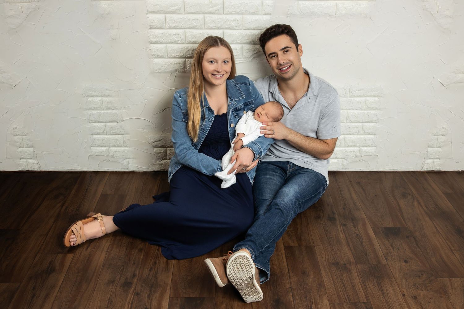Happy young couple sitting on hardwood floor against white brick wall, smiling and holding their newborn baby.
