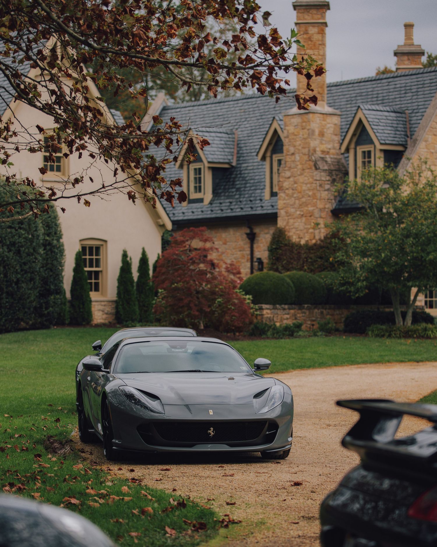 Gray Ferrari 812 parked in front of a luxurious stone manor house surrounded by lush gardens.