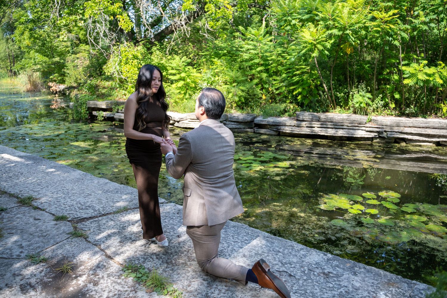 Man proposes on one knee to woman by a serene pond surrounded by lush greenery in an outdoor setting.