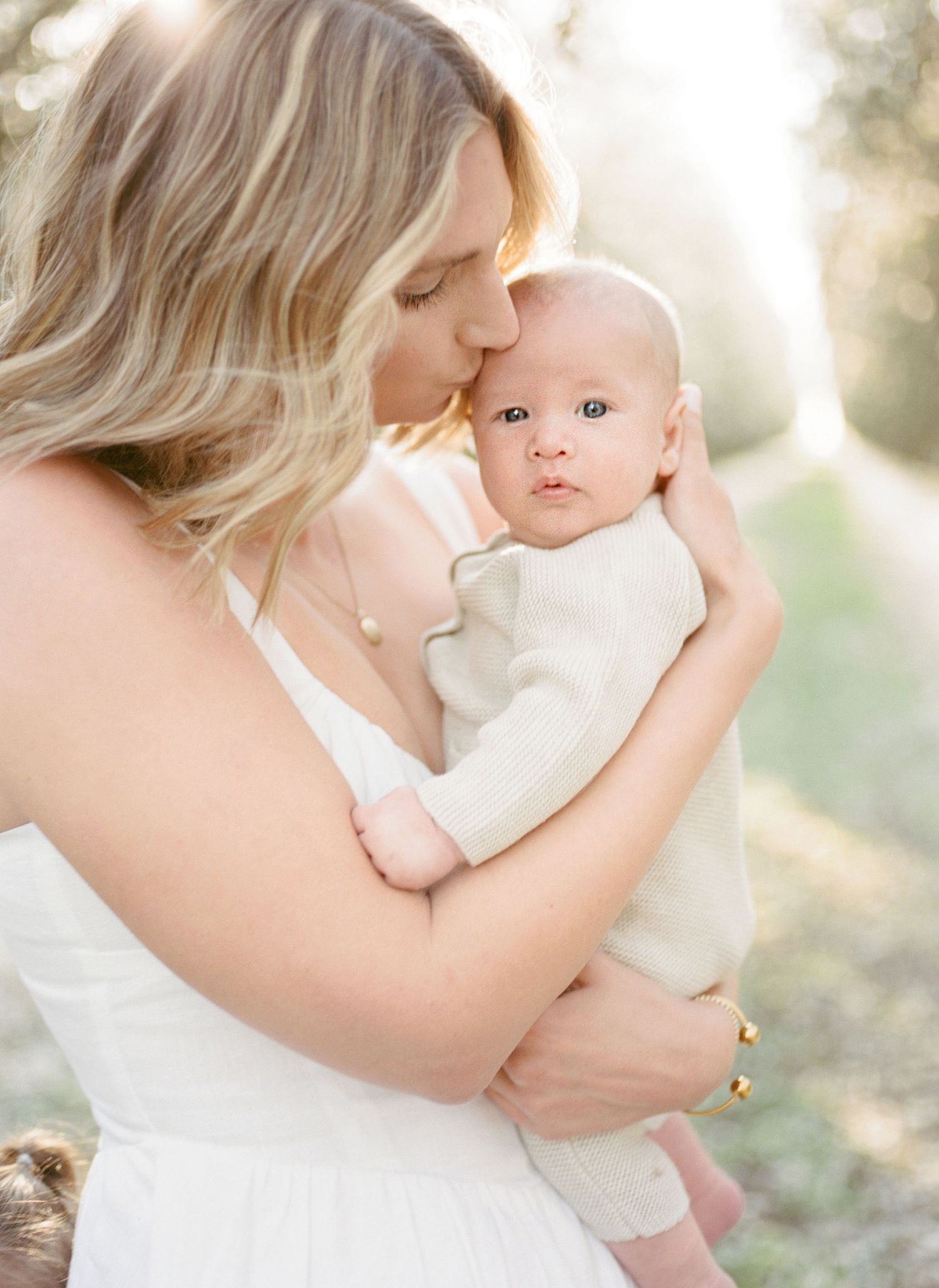 A mother in a white dress tenderly kisses her newborn baby outdoors in soft golden sunlight.
