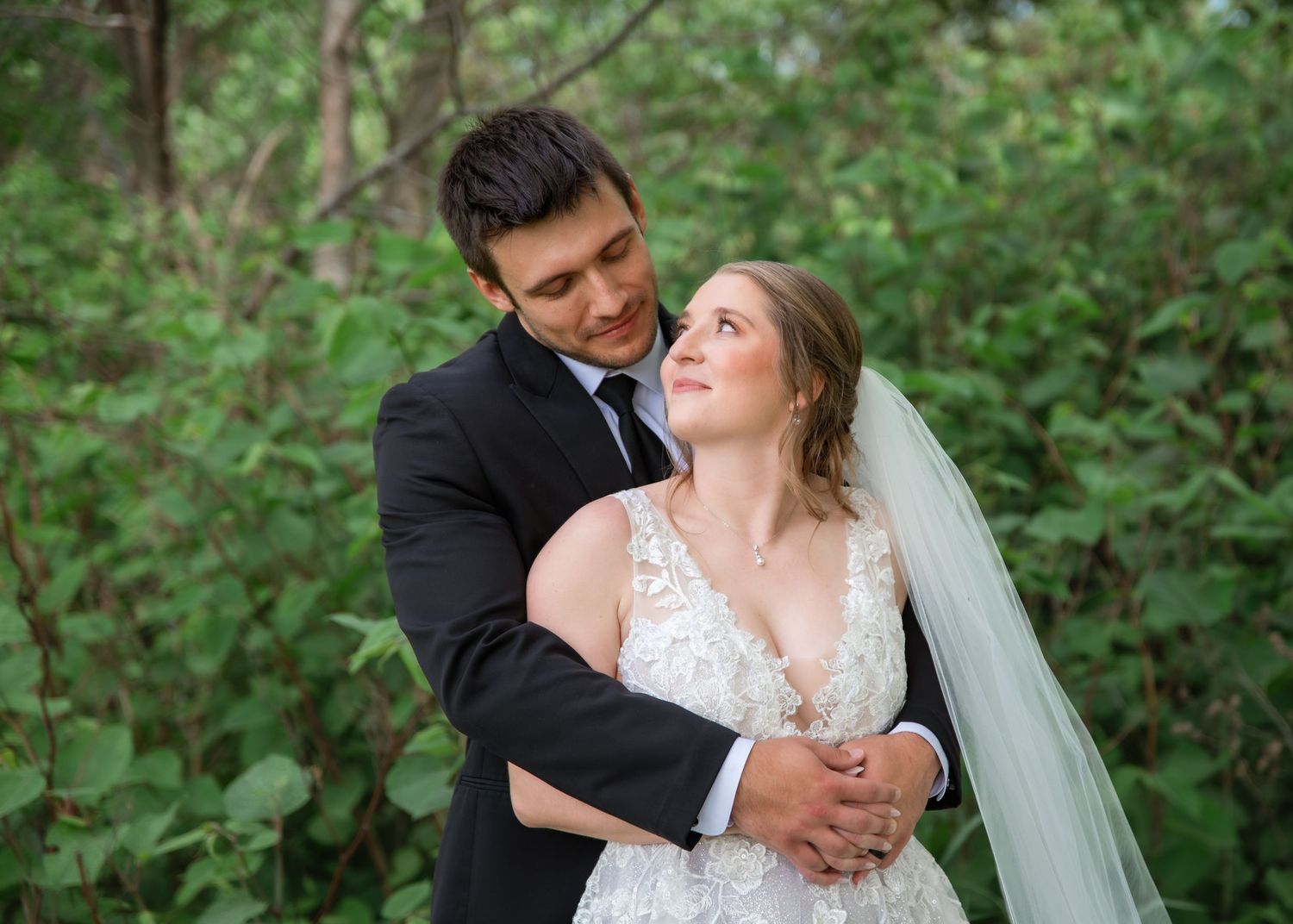 Bride and groom embrace outdoors among lush green foliage, bride in lace gown with veil gazing up lovingly.