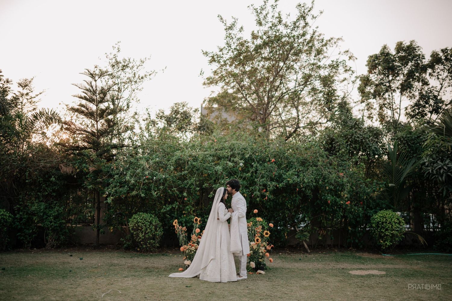Couple in white wedding attire sharing a romantic moment in a lush green garden at sunset.
