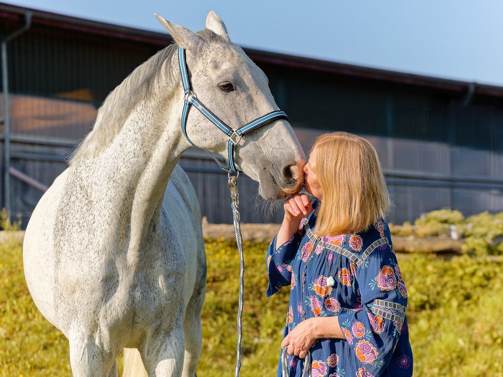 Frau in blauer Bluse mit Schimmel vor Reithalle. Professionelle Outdoor-Tierporträts von Lichtkapsel in Wiesenttal.