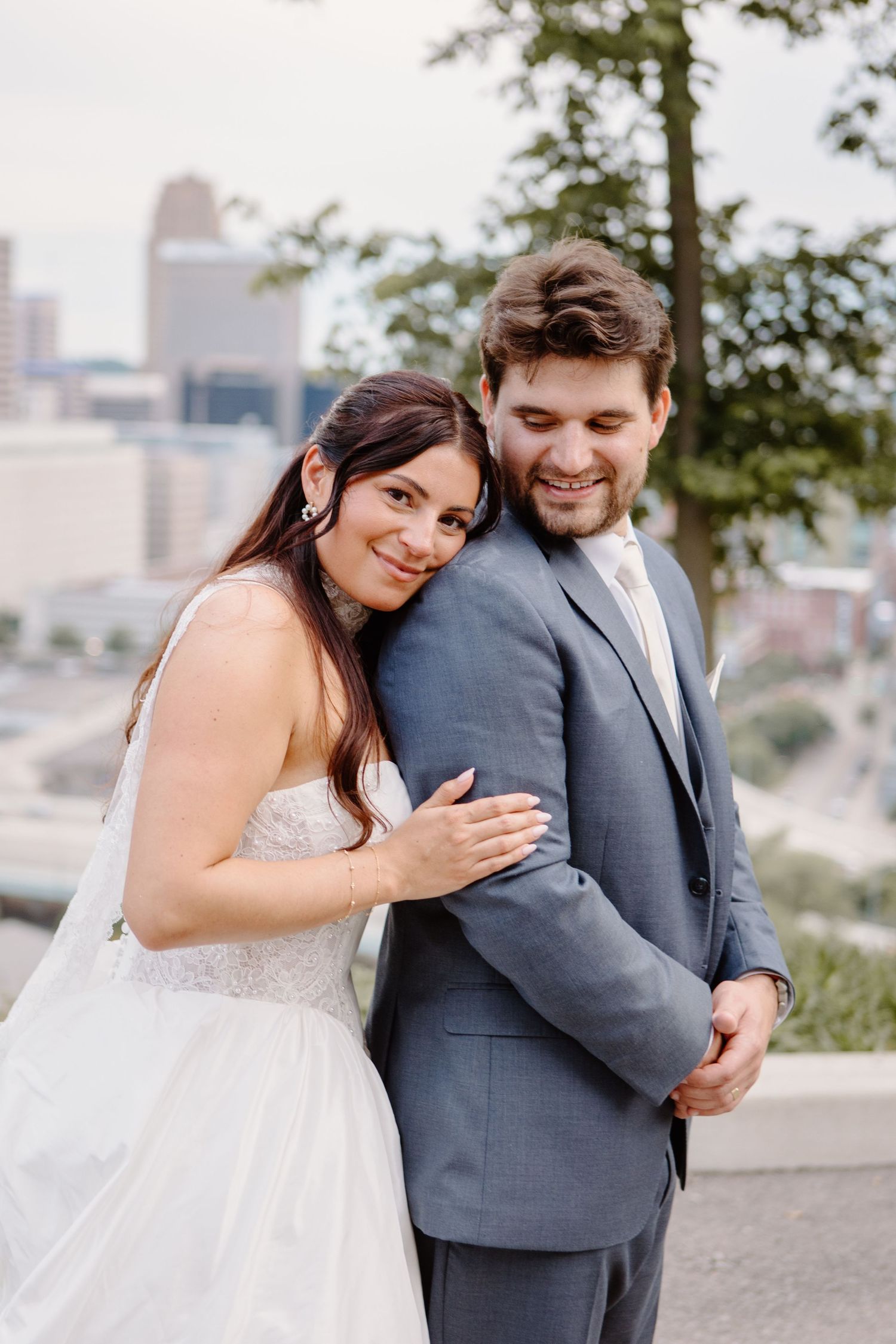 Bride in white gown leans on groom in gray suit, smiling outdoors with city skyline in background.