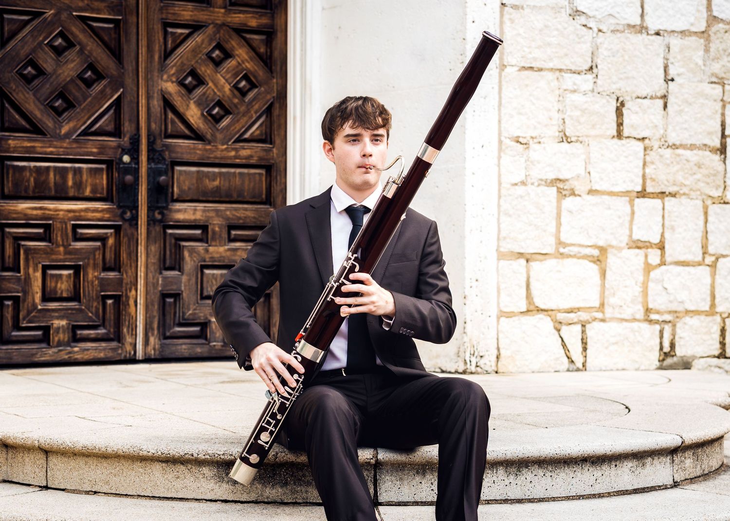 Young male musician in a suit holding a bassoon, seated on steps near a wooden door and stone wall.