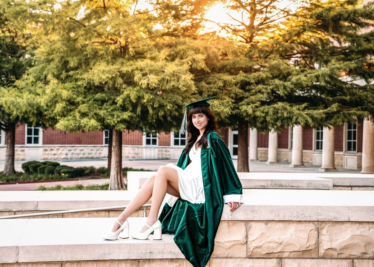 A smiling graduate in green cap and gown sits on a stone wall outdoors on a sunny college campus.