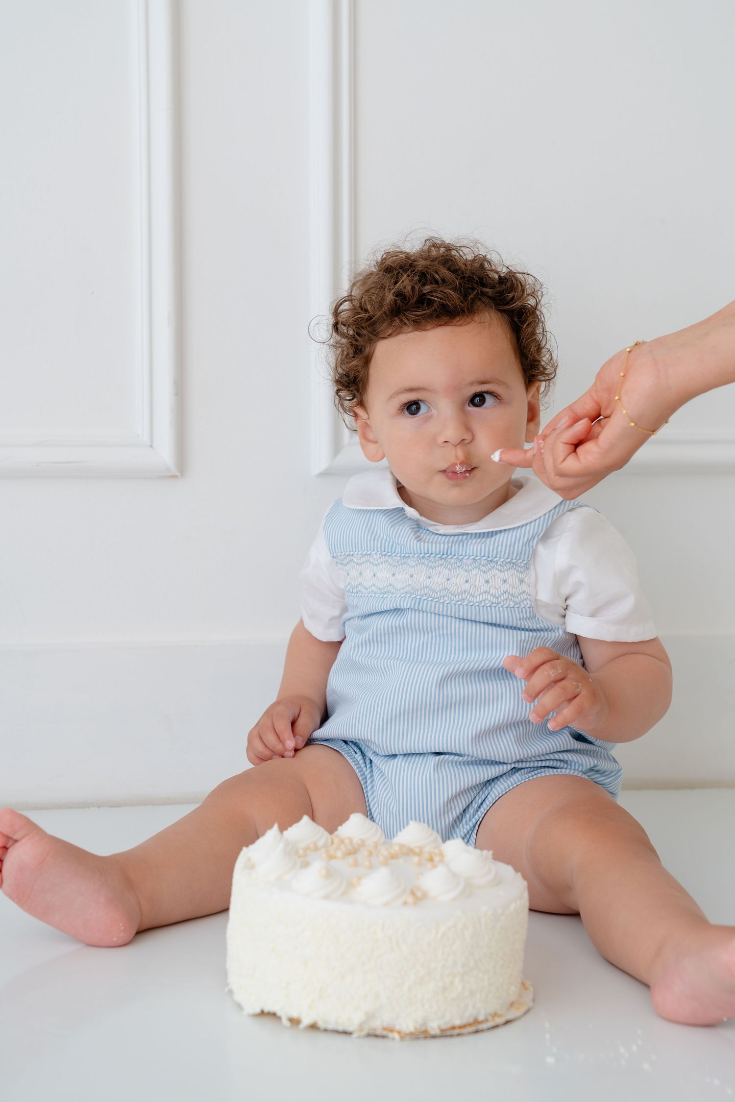 Curly-haired baby in blue romper sits by white cake, being fed frosting by adult hand during first birthday celebration.