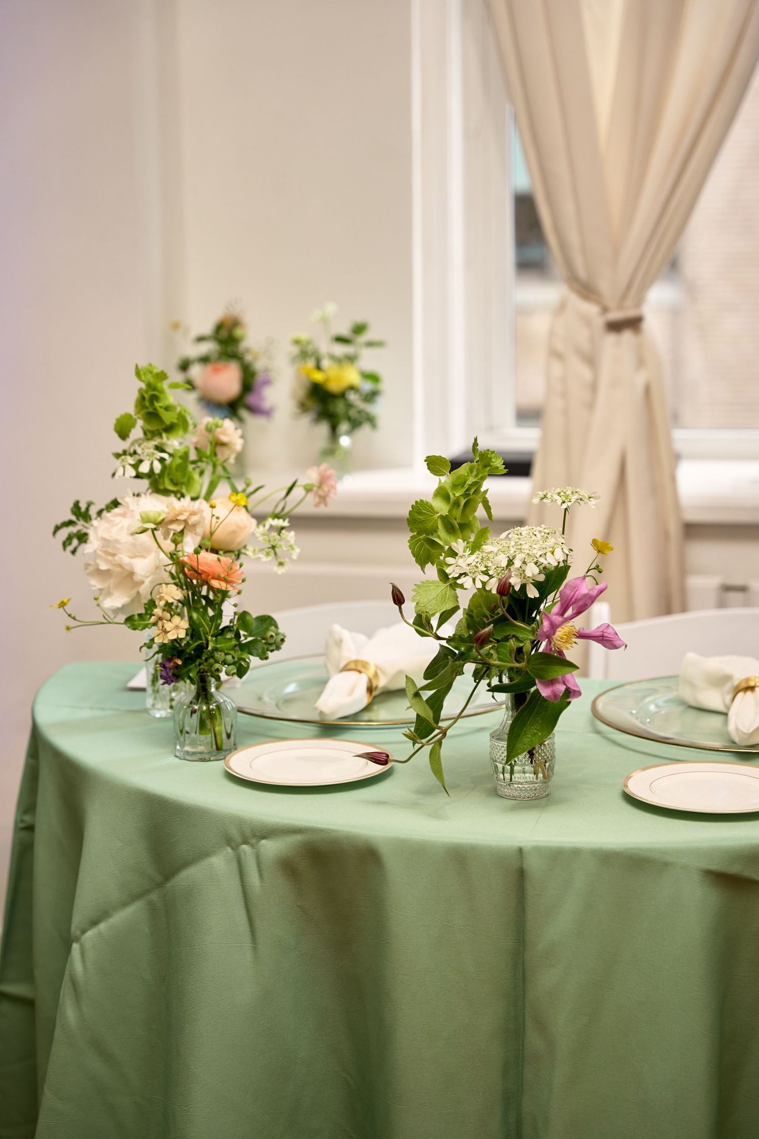 Elegant table setting with green tablecloth, floral centerpieces, and plates in a bright event venue.
