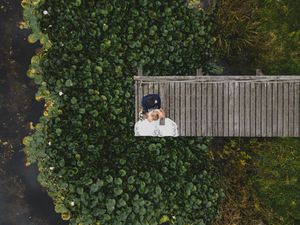 Aerial view of a person lying on a wooden boardwalk surrounded by lush green water plants near a pond.