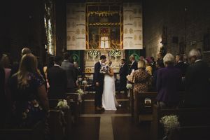 Bride and groom exchange vows at a candlelit stone church altar, surrounded by seated wedding guests in wooden pews.