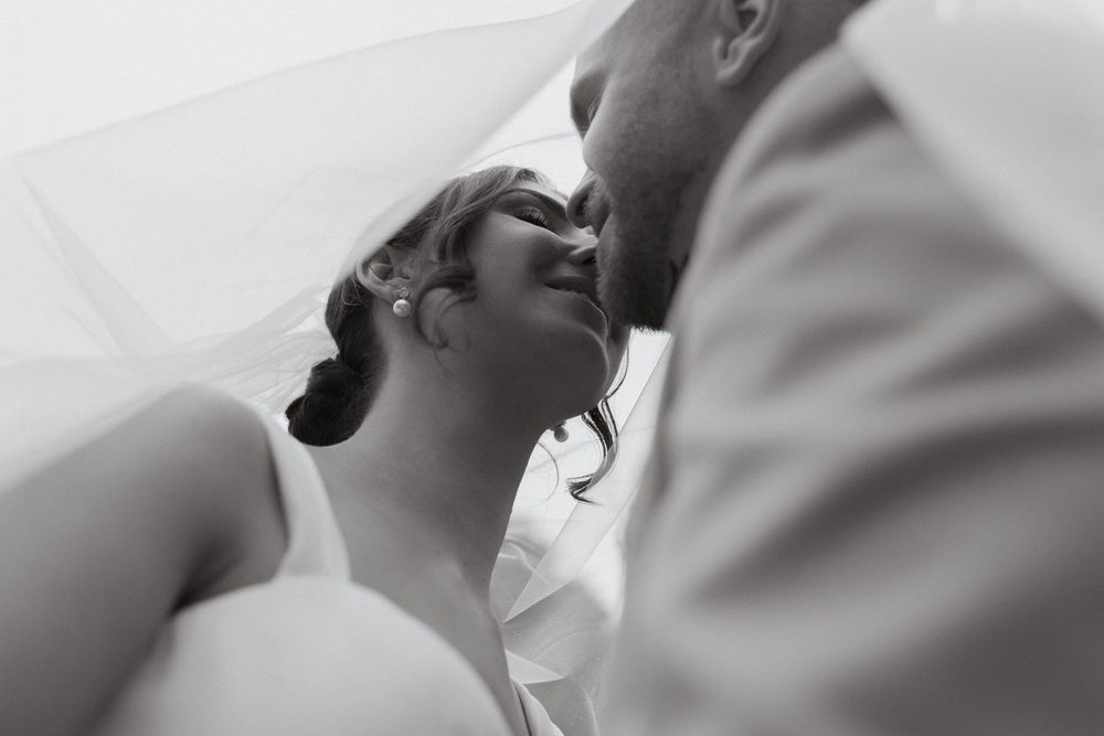 Black and white photo of a bride and groom sharing a tender kiss beneath a flowing wedding veil.