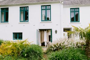 White two-story house with green-trimmed windows, open shutters displaying a wedding dress, surrounded by lush garden greenery.