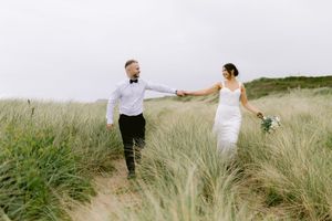 A bride and groom walk hand-in-hand through tall grass dunes on a beach, with the bride holding a bouquet.