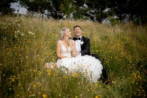 Bride and groom sitting together laughing in a wildflower meadow, bride in white gown, groom in black tuxedo.