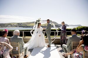 Outdoor wedding ceremony on a terrace with ocean views, bride in white gown and groom in gray suit exchanging vows.