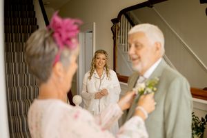 Bride in white robe smiles at elderly couple adjusting boutonniere near staircase during wedding preparations.