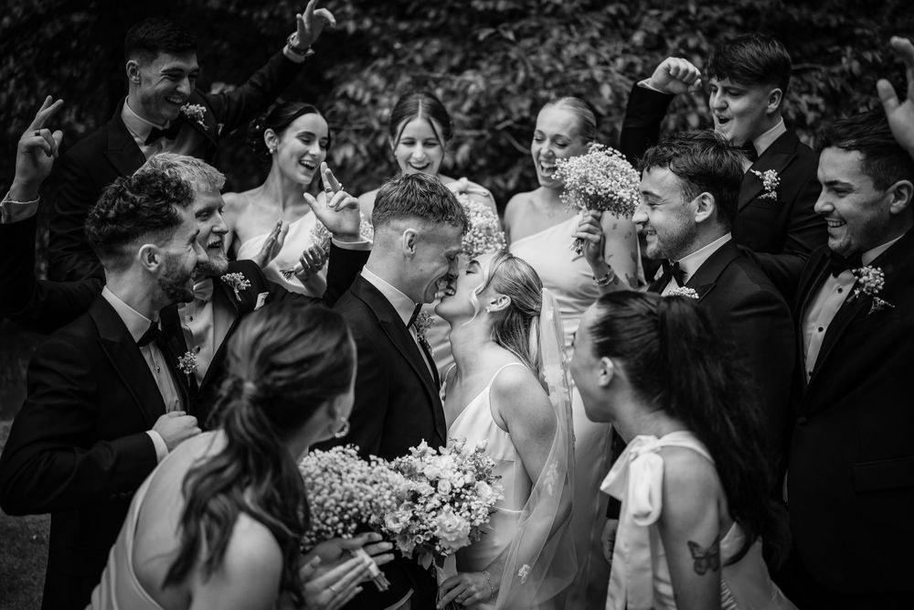 Black and white wedding photo of bride and groom kissing surrounded by cheering bridal party outdoors.