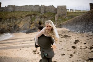 Couple enjoying a piggyback ride on a sandy beach with a historic castle ruin in the background.